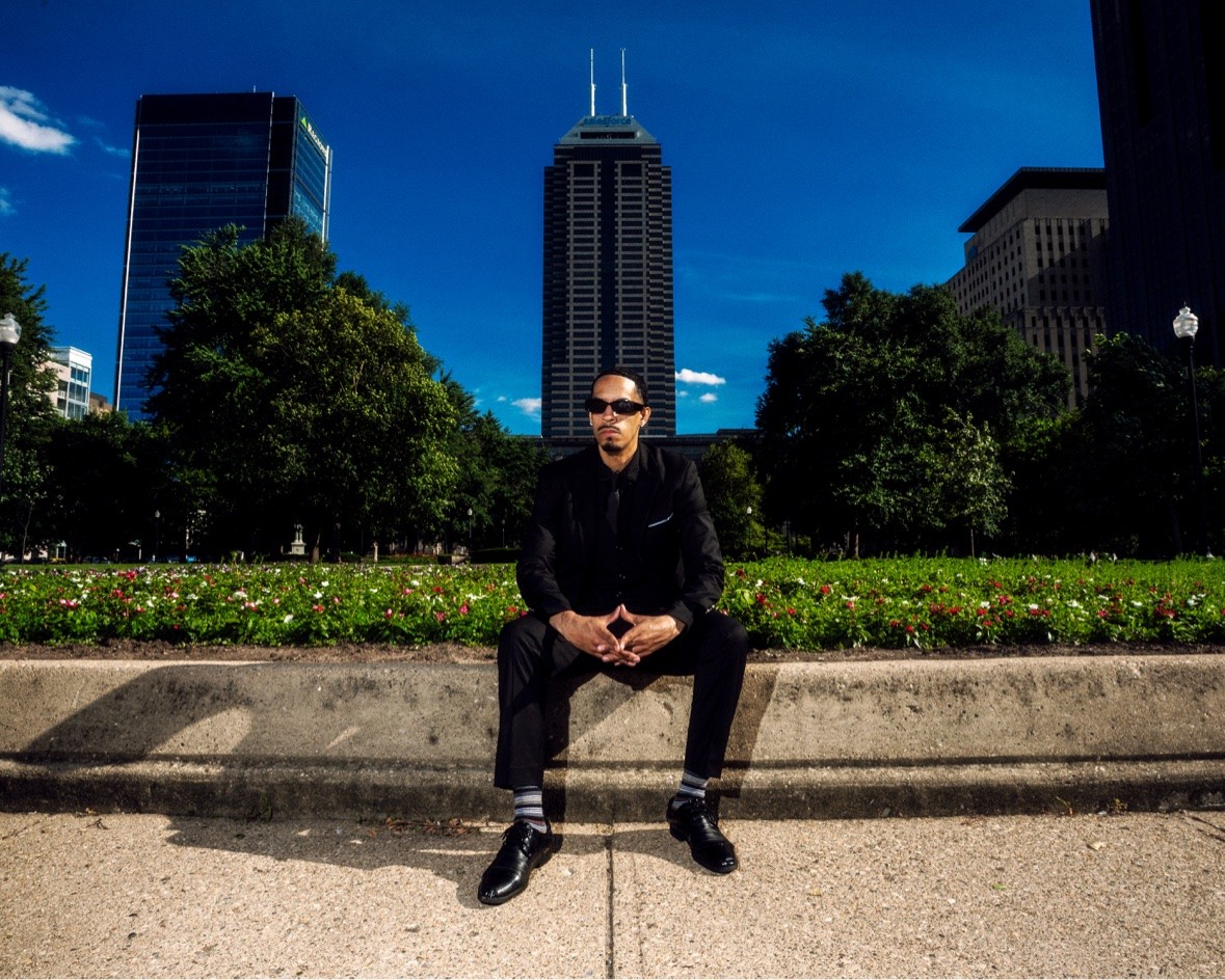 Black suit seated in a park with the city skyline behind him